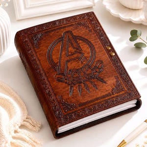 May include: A brown leather-bound journal featuring an embossed Avengers logo on the front cover. The cover has intricate decorative border patterns and two small metallic studs on the side. The journal contains white paper pages and has a rectangular shape.