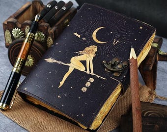 May include: A dark blue leather journal with a witch riding a broomstick design, a crescent moon, and stars. The journal is open, revealing aged paper. A wooden pencil and a pen with gold accents are next to the journal.