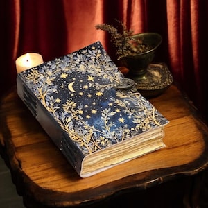 May include: A decorative book with a celestial design. The cover features a dark blue background with a crescent moon and stars, surrounded by gold and white floral patterns. The book is secured with a metal clasp and rests on a wooden surface.