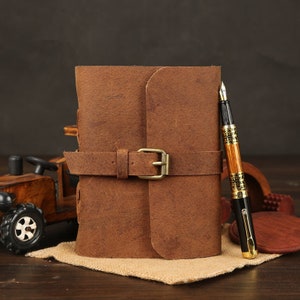 May include: A brown leather journal with a buckle closure and a gold-toned pen on a wooden surface.