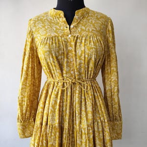 Könnte beinhalten: Senfgelbes Kleid mit weißem Blumenmuster. Das langärmlige Kleid hat eine Knopfleiste vorne, eine betonte Taille mit passendem Gürtel und einen Stufenrock. Das Kleid wird auf einer Schaufensterpuppe präsentiert.