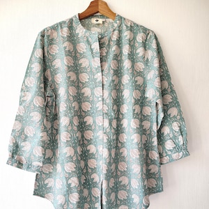 May include: Teal blouse with a floral design, mandarin collar, and three-quarter sleeves. The shirt is fastened with buttons and hangs from a wooden hanger. The floral pattern is in a lighter shade.