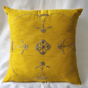 May include: Mustard yellow decorative pillow with embroidered floral and sequin design. The square pillow features a central floral motif and a border of delicate silver sequins, adding a touch of elegance to home decor.
