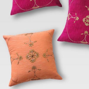 May include: Close-up of decorative throw pillows in shades of pink and peach. The pillows feature intricate gold floral embroidery and embellishments. The pillows are square-shaped and made of a textured fabric.
