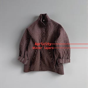 Puede incluir: Chaqueta acolchada marrón oscuro con líneas de puntos blancos verticales. La chaqueta tiene cuello alto y mangas enrolladas. El texto "Apricity Winter layers" es visible.