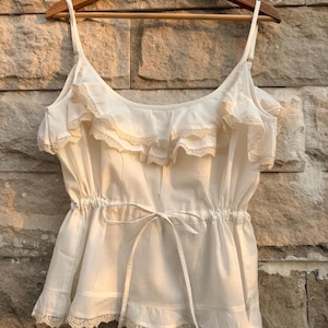 May include: A white camisole top with spaghetti straps, a ruffled neckline, and a drawstring waist. The top features delicate lace trim along the neckline and hem. The garment is hanging on a wooden hanger against a stone wall.