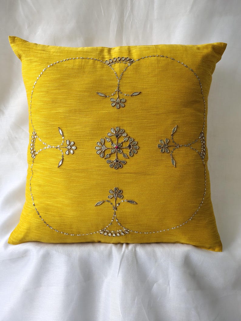 May include: Mustard yellow decorative pillow with embroidered floral and sequin design. The square pillow features a central floral motif and a border of delicate silver sequins, adding a touch of elegance to home decor.