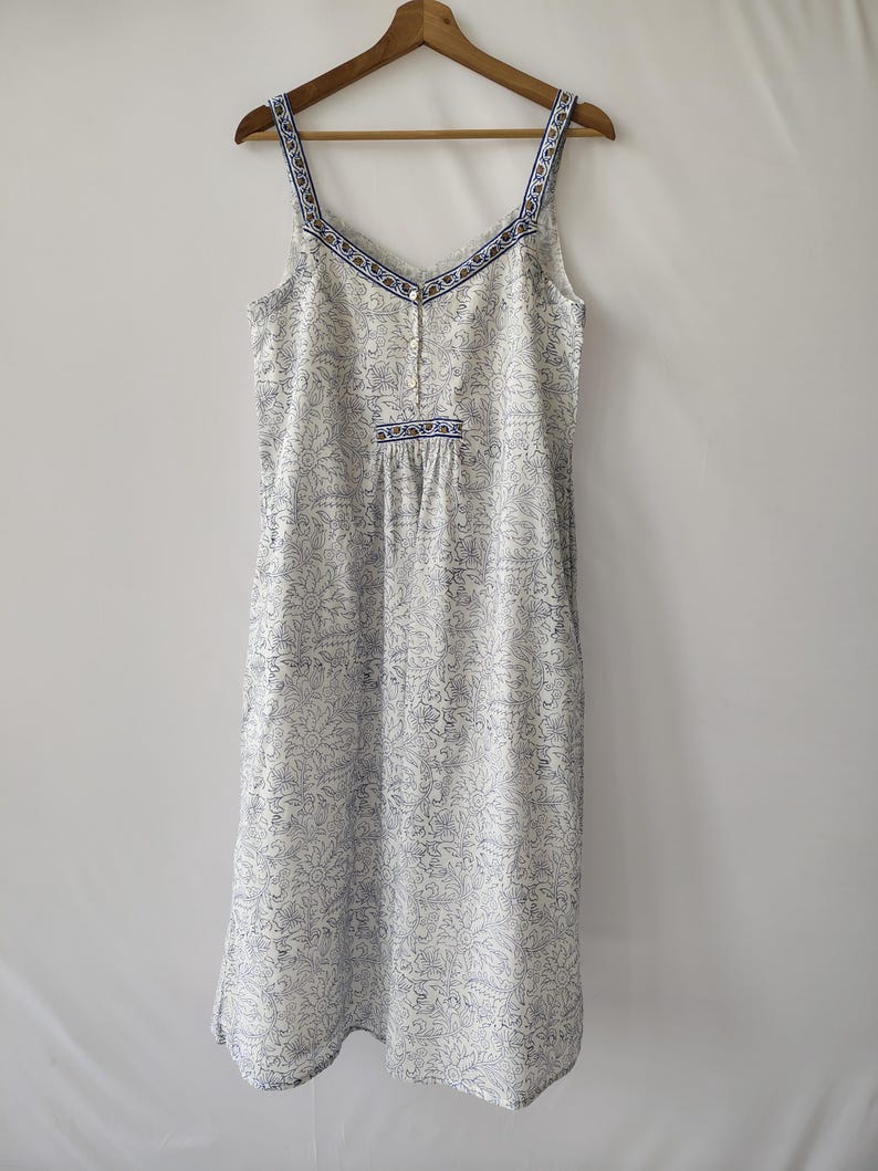 Puede incluir: Camis&oacute;n blanco con estampado floral azul, colgado de una percha de madera. El escote y los tirantes est&aacute;n adornados con un dise&ntilde;o azul y blanco. El camis&oacute;n tiene cuello en V y tirantes finos.