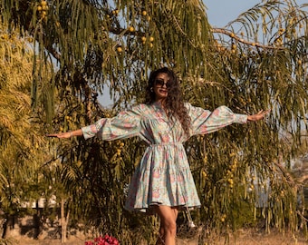 Può includere: Una donna con un vestito floreale azzurro chiaro con maniche a sbuffo si trova in un campo di erba secca. Sorride e guarda la telecamera. In primo piano c'è una borsa marrone e dei fiori rossi.