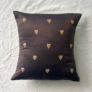 May include: A square, dark brown decorative pillow with gold-colored embroidered embellishments. The pillow has a silky texture and is adorned with repeating, stylized floral or leaf-like designs.