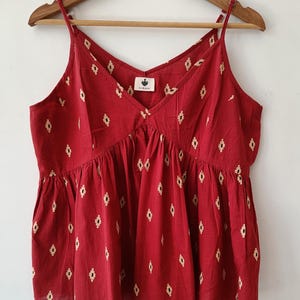 Puede incluir: Un top rojo sin mangas con cuello en V y un patrón geométrico blanco. El top tiene un dobladillo con volantes y una pequeña etiqueta que dice "SUKHI".