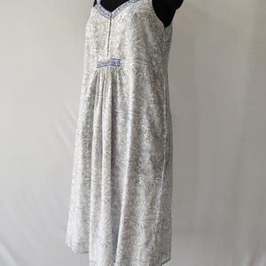 Puede incluir: Vestido blanco y azul con estampado floral, escote en V y tirantes finos. El vestido presenta una cintura fruncida y un dobladillo a la rodilla. Los tirantes y el escote est&aacute;n ribeteados con un borde azul. El vestido se exhibe en un maniqu&iacute; negro.