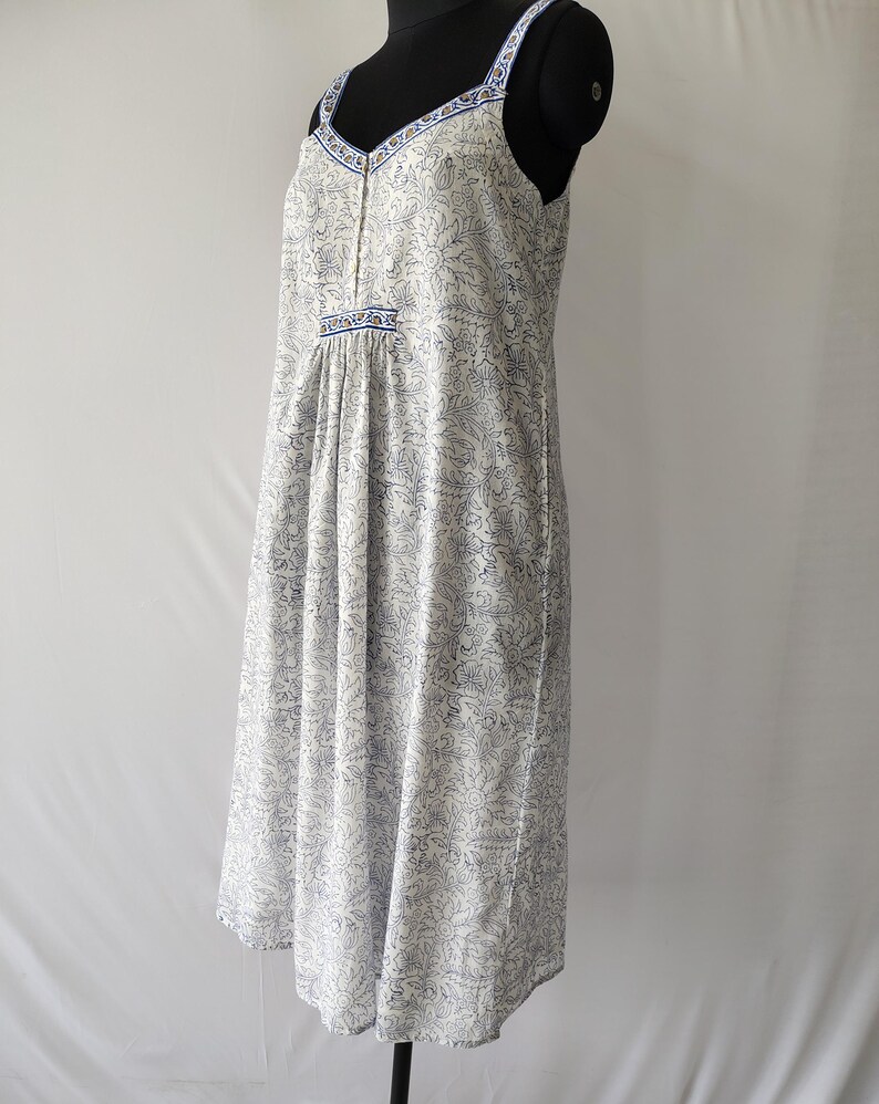 Puede incluir: Vestido blanco y azul con estampado floral, escote en V y tirantes finos. El vestido presenta una cintura fruncida y un dobladillo a la rodilla. Los tirantes y el escote est&aacute;n ribeteados con un borde azul. El vestido se exhibe en un maniqu&iacute; negro.