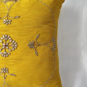 May include: A square, mustard-yellow decorative pillow with intricate embroidery. The embroidery features floral and leaf patterns in silver thread, with small sequins adding sparkle. The pillow is made of a textured fabric.