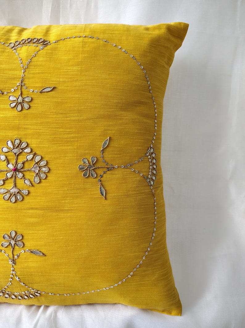 May include: A square, mustard-yellow decorative pillow with intricate embroidery. The embroidery features floral and leaf patterns in silver thread, with small sequins adding sparkle. The pillow is made of a textured fabric.