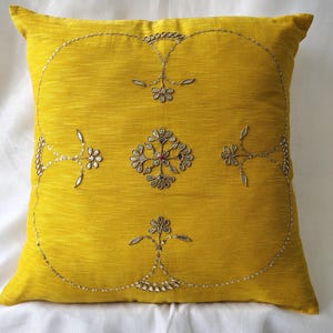 May include: A square, yellow decorative pillow with intricate silver embroidery. The embroidery features floral and geometric patterns, with a central floral design and a beaded border. The pillow is likely made of a textured fabric.
