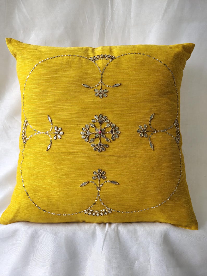 May include: A square, yellow decorative pillow with intricate silver embroidery. The embroidery features floral and geometric patterns, with a central floral design and a beaded border. The pillow is likely made of a textured fabric.