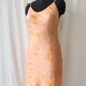 May include: Peach-colored slip dress with a tie-shoulder design. The dress features a subtle tie-dye pattern in shades of yellow and orange. The dress is displayed on a black mannequin.