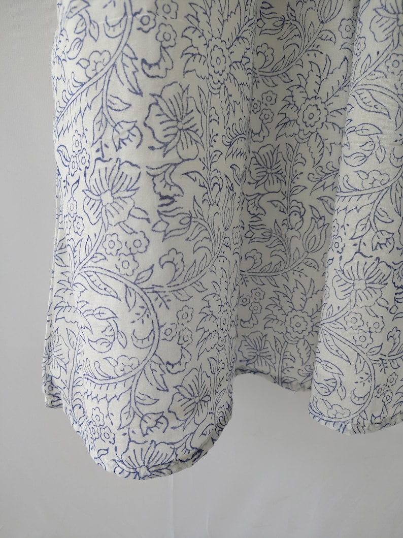 Puede incluir: Primer plano de una tela blanca con un patr&oacute;n floral azul repetido. El dise&ntilde;o presenta flores, hojas y enredaderas estilizadas. La tela parece estar drapeada, con pliegues y curvas suaves.