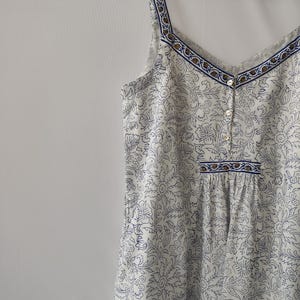 Puede incluir: Vestido camisola blanco con estampado floral azul, cuello en V y detalles de botones. El vestido presenta un ribete con estampado azul y marr&oacute;n a lo largo del escote y los tirantes. Colgado en una percha de madera.