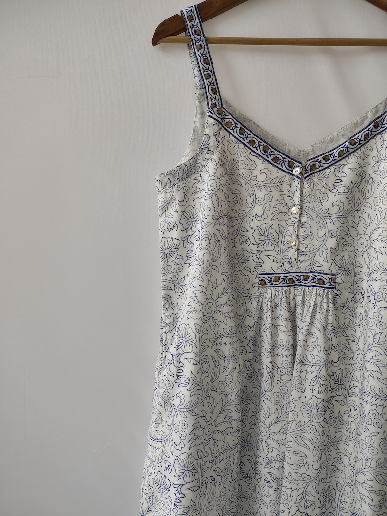 Puede incluir: Vestido camisola blanco con estampado floral azul, cuello en V y detalles de botones. El vestido presenta un ribete con estampado azul y marr&oacute;n a lo largo del escote y los tirantes. Colgado en una percha de madera.