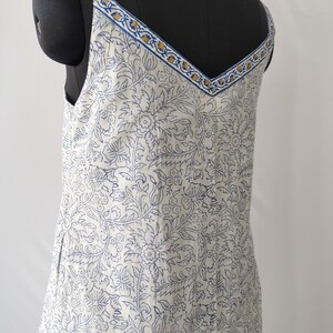 Puede incluir: Vestido blanco con estampado floral azul, con escote en V y tirantes finos. Los tirantes y el escote est&aacute;n adornados con un dise&ntilde;o azul y dorado. El vestido se muestra en un maniqu&iacute; negro.