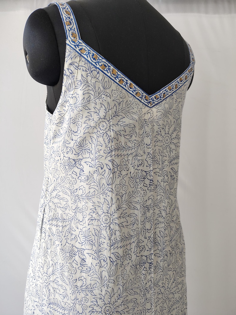 Puede incluir: Vestido blanco con estampado floral azul, con escote en V y tirantes finos. Los tirantes y el escote est&aacute;n adornados con un dise&ntilde;o azul y dorado. El vestido se muestra en un maniqu&iacute; negro.