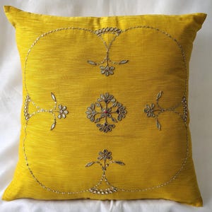 May include: A square, yellow decorative pillow with an embroidered floral design. The embroidery features silver-colored thread and sequins, creating a detailed pattern. The pillow is a solid color with a textured appearance.