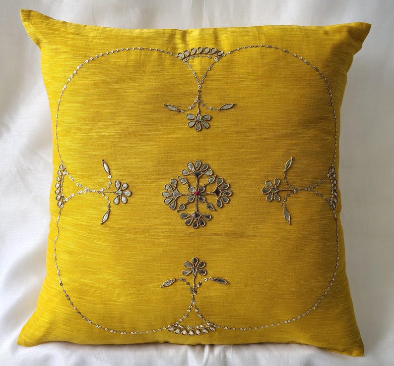 May include: A square, yellow decorative pillow with an embroidered floral design. The embroidery features silver-colored thread and sequins, creating a detailed pattern. The pillow is a solid color with a textured appearance.