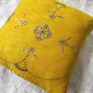 May include: A square, yellow decorative pillow with intricate silver embroidery. The embroidery features floral and geometric patterns, with a central design and delicate embellishments. The pillow is made of a textured fabric.