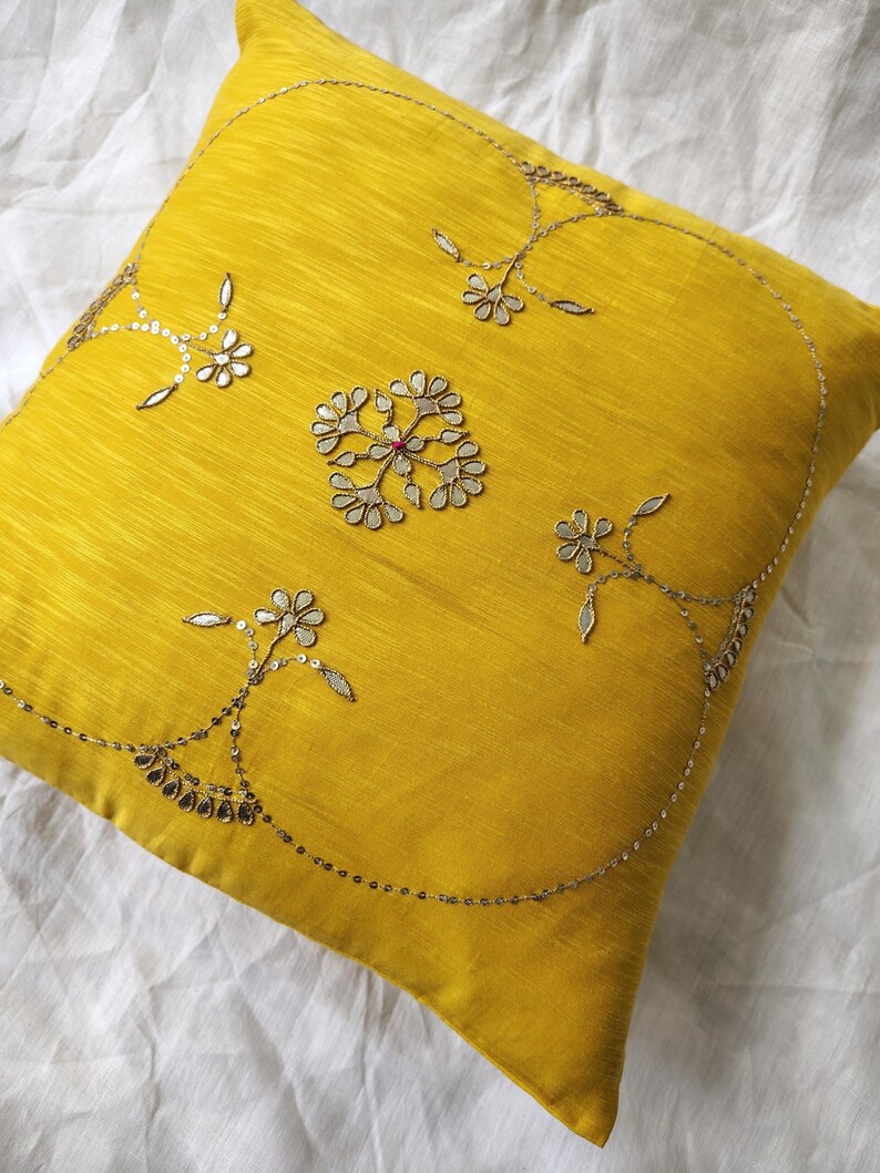May include: A square, yellow decorative pillow with intricate silver embroidery. The embroidery features floral and geometric patterns, with a central design and delicate embellishments. The pillow is made of a textured fabric.