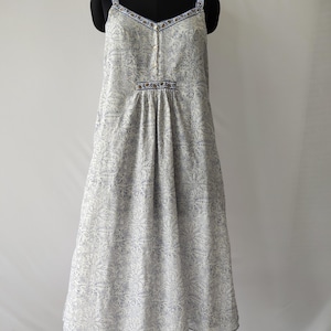 Puede incluir: Vestido blanco con estampado floral azul, con tirantes finos y escote en V. El vestido presenta una botonadura frontal y una cintura fruncida. La tela tiene un delicado estampado floral en azul, y los tirantes y el escote est&aacute;n ribeteados en azul.