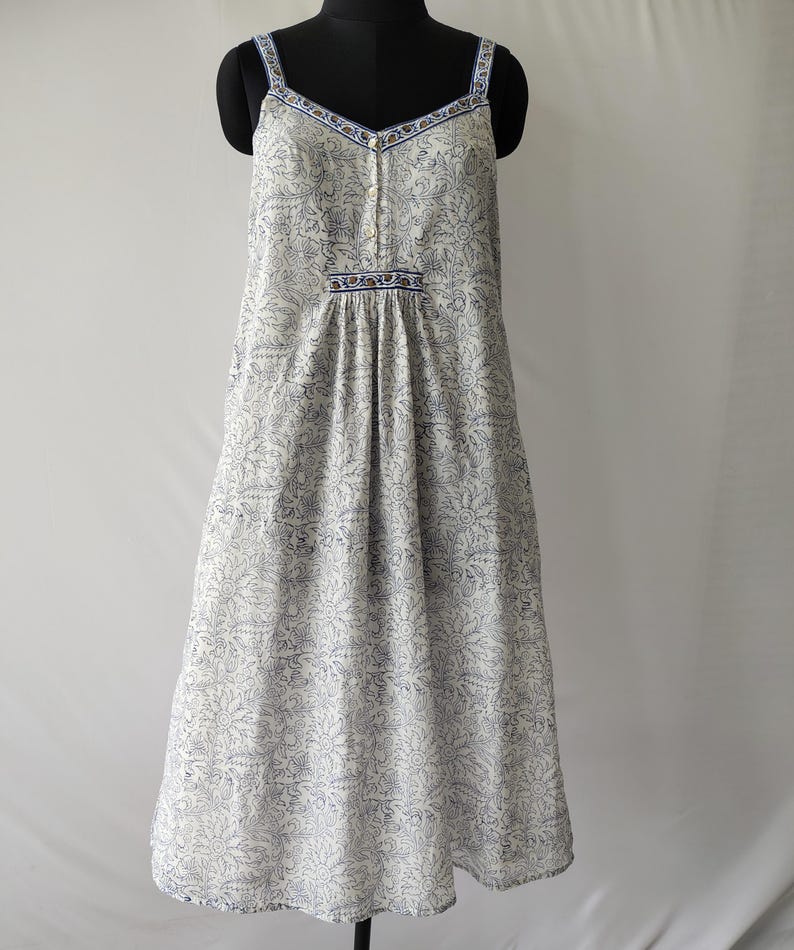 Puede incluir: Vestido blanco con estampado floral azul, con tirantes finos y escote en V. El vestido presenta una botonadura frontal y una cintura fruncida. La tela tiene un delicado estampado floral en azul, y los tirantes y el escote est&aacute;n ribeteados en azul.