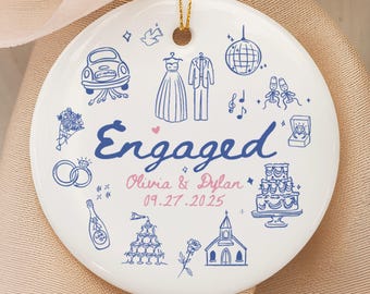 May include: A white ceramic ornament with a gold string, featuring blue and pink illustrations of wedding-related icons. The word "Engaged" is in blue script, with names and a date below.