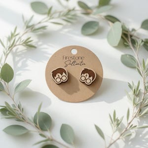 May include: Wooden stud earrings featuring a cartoon character with round glasses and a lightning bolt scar. The earrings are on a brown card with the text "Firestone Saltwater". The earrings are surrounded by green leaves.