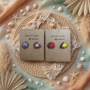 May include: Two pairs of handmade stud earrings on display. One pair features a seashell and a cartoon character, the other pair features a crab and a smiling yellow character. Each pair is on a card that reads "Handmade with love."