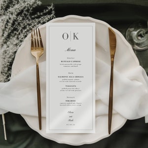 May include: A white menu card with black text and a gold border. The menu card has the initials "OK" at the top and the word "Menu" below. The menu lists the following items: Buffalo Caprese, Salmone Alla Griglia, Tagliata, and Tiramisu. The menu card is on a white plate with a gold fork and knife.