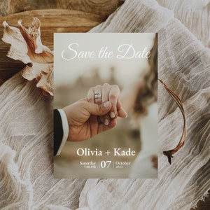 Op de afbeelding: Save the Date kaart met de handen van een stel dat elkaar vasthoudt, met een ring aan de vinger van de vrouw. De kaart ligt op een rustieke houten achtergrond met een stuk stof eroverheen gelegd. De tekst op de kaart luidt "Save the Date" in een cursief schrift, gevolgd door "Olivia + Kade" en "Zaterdag 07 oktober 2023".