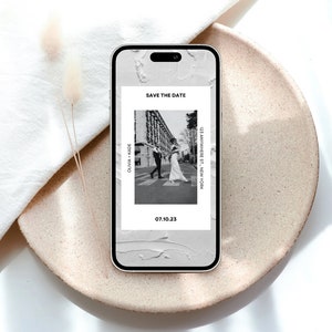 Könnte beinhalten: Ein Smartphone-Bildschirm zeigt eine Save-the-Date-Ankündigung mit einem Schwarzweißfoto eines Paares, das eine Stadtstraße entlangläuft. Der Text lautet "Save the Date" und "Olivia + Jade" mit dem Datum "07.10.23" und dem Ort "123 Anywhere St., New York".