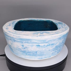 Pode incluir: Uma tigela de cerâmica artesanal com interior retangular. O exterior é pintado com um design de pinceladas em azul claro e branco, e o interior é de cor verde-azulado escuro. A tigela é exibida em um suporte giratório branco e preto.