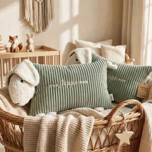 May include: Two rectangular sage green corduroy pillows shaped like sheep with cream-colored textured fleece heads and ears. The pillows feature the embroidered name Jeppe flanked by small heart icons. They are nestled in a woven wicker basket.