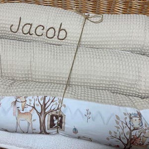 May include: A beige waffle-textured baby nest with the name Jacob embroidered in brown thread. The outer border features a white fabric with a printed pattern of forest animals, including deer and owls, and trees. A thin brown string ties the sides.