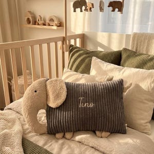 Peut inclure: Un coussin rectangulaire en velours c&ocirc;tel&eacute; gris fonc&eacute; en forme d'&eacute;l&eacute;phant avec une t&ecirc;te et des oreilles en peluche cr&egrave;me. Le coussin pr&eacute;sente le nom Tino brod&eacute; en &eacute;criture blanche au centre de la section en velours c&ocirc;tel&eacute; gris fonc&eacute;.