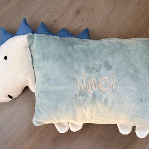 May include: A plush dinosaur-shaped pillow in light blue and white. The pillow features a white head with black eyes and blue spikes along the back. The word "Noel" is embroidered in a cursive font on the pillow's body.