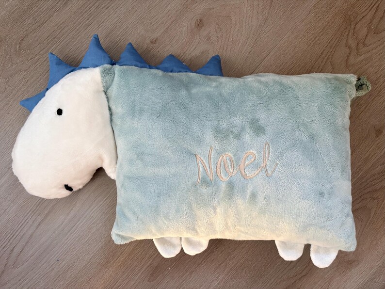 May include: A plush dinosaur-shaped pillow in light blue and white. The pillow features a white head with black eyes and blue spikes along the back. The word "Noel" is embroidered in a cursive font on the pillow's body.