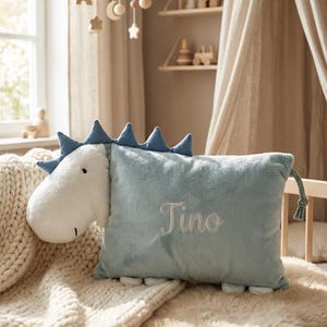May include: A plush dinosaur-shaped pillow in light blue and white, with the name "Tino" embroidered on the front. The pillow has blue spikes and is placed on a bed with a cream-colored blanket and a faux fur throw.
