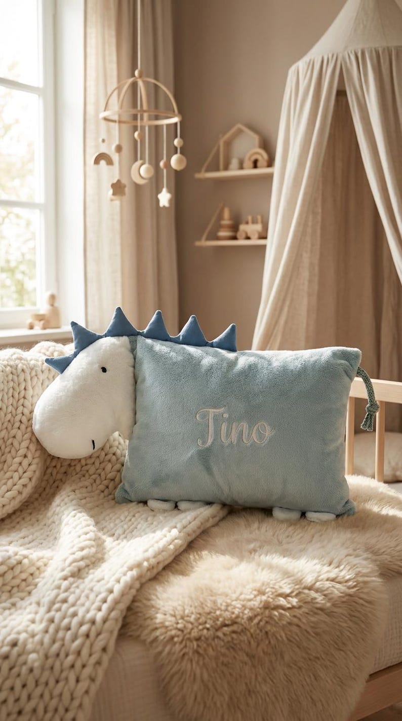 May include: A plush dinosaur-shaped pillow in light blue and white, with the name "Tino" embroidered on the front. The pillow has blue spikes and is placed on a bed with a cream-colored blanket and a faux fur throw.