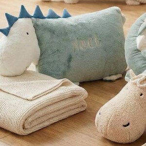 May include: A collection of plush dinosaur-themed nursery items. Includes a white and blue dinosaur head, a light blue pillow with "Noel" embroidered, a folded beige blanket, and a plush hippo.
