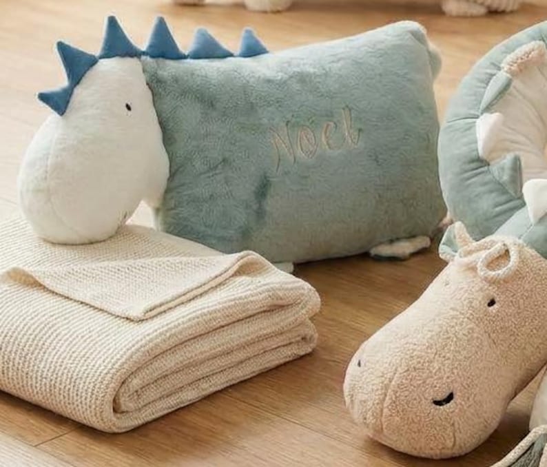 May include: A collection of plush dinosaur-themed nursery items. Includes a white and blue dinosaur head, a light blue pillow with "Noel" embroidered, a folded beige blanket, and a plush hippo.