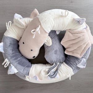 Op de afbeelding: Een cirkelvormig babyspeelgoed in crème, grijs en beige, met een paardenkop. Het pluche speelgoed heeft een zacht, getextureerd lichaam met zwarte stipogen. Het bevat linten en verschillende texturen voor sensorisch spel.